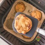 A golden crispy chocolate chip cookie on spatula coming up from air fryer basket with four other cookies.