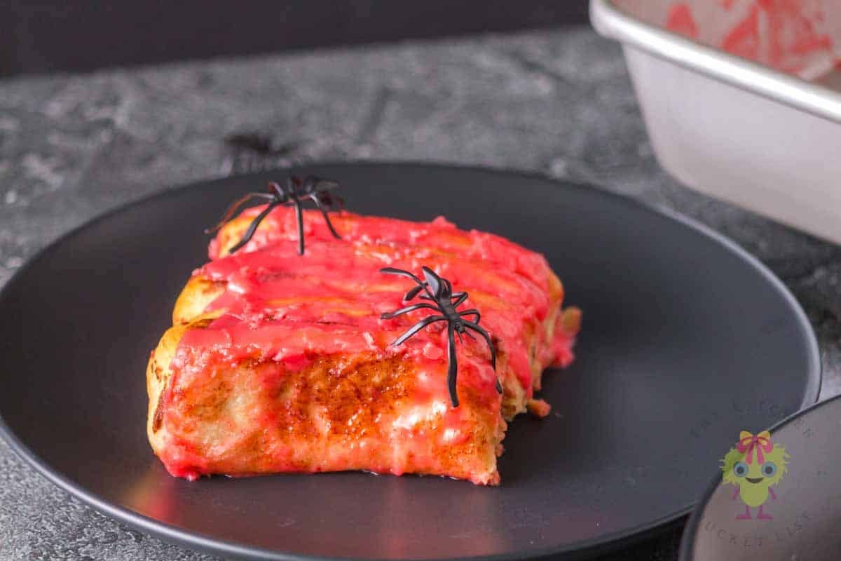 Halloween cinnamon rolls shaped like intestines on a black plate with toy spiders. The red icing adds the perfect touch of gore.
