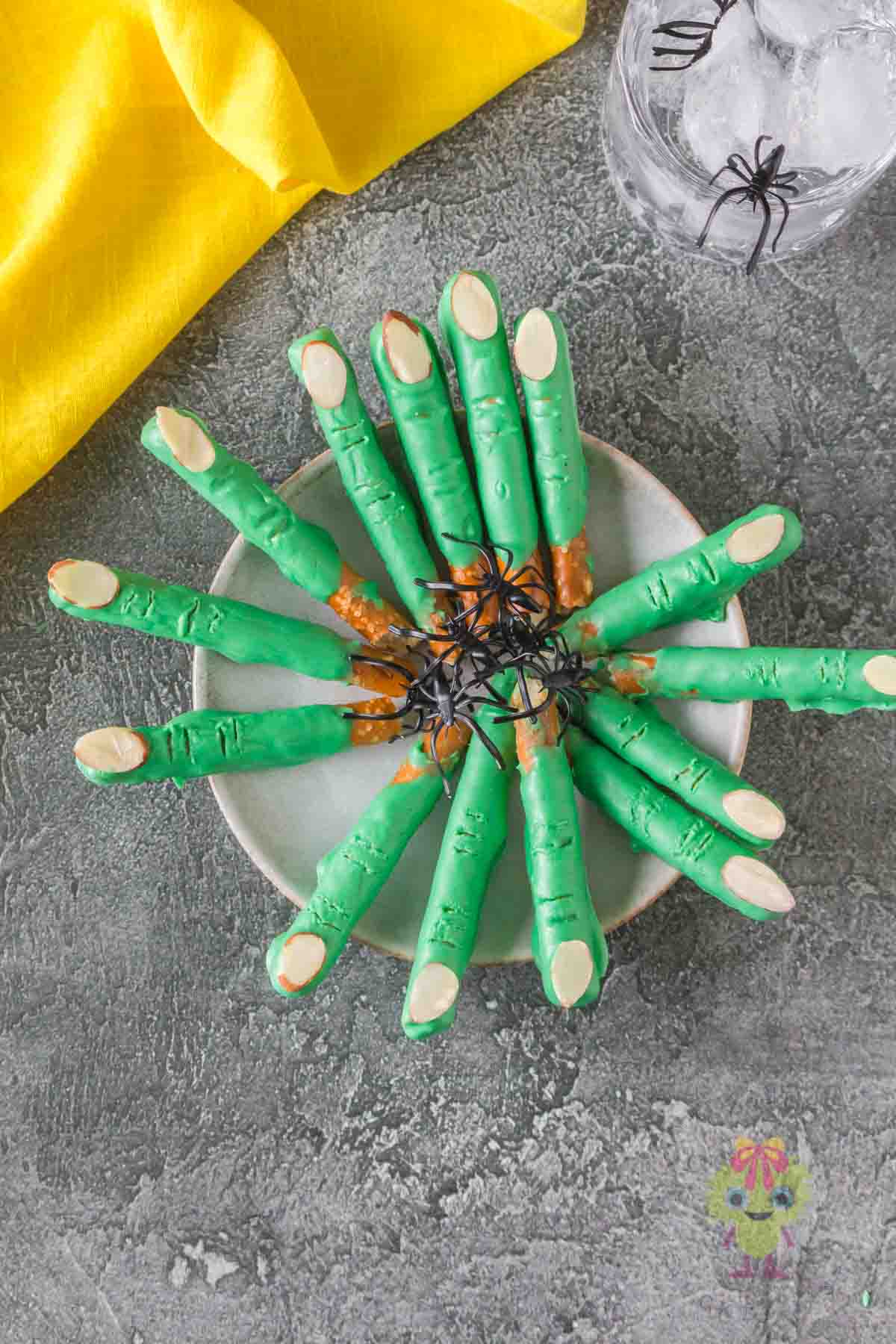 Halloween pretzels decorated and shaped into witch fingers pretzels on a serving plate with toy spiders in the middle.
