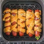 Overhead shot of the piggies in a blanket in the air fryer basket. They are done up for Halloween to look like mummy dogs with googly eyes.