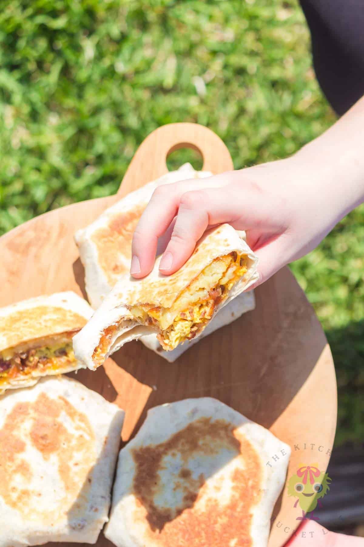 Four Taco Bell breakfast crunchwraps on a wooden cutting board, with one cut in half. One half is held above the others with a bite taken out.