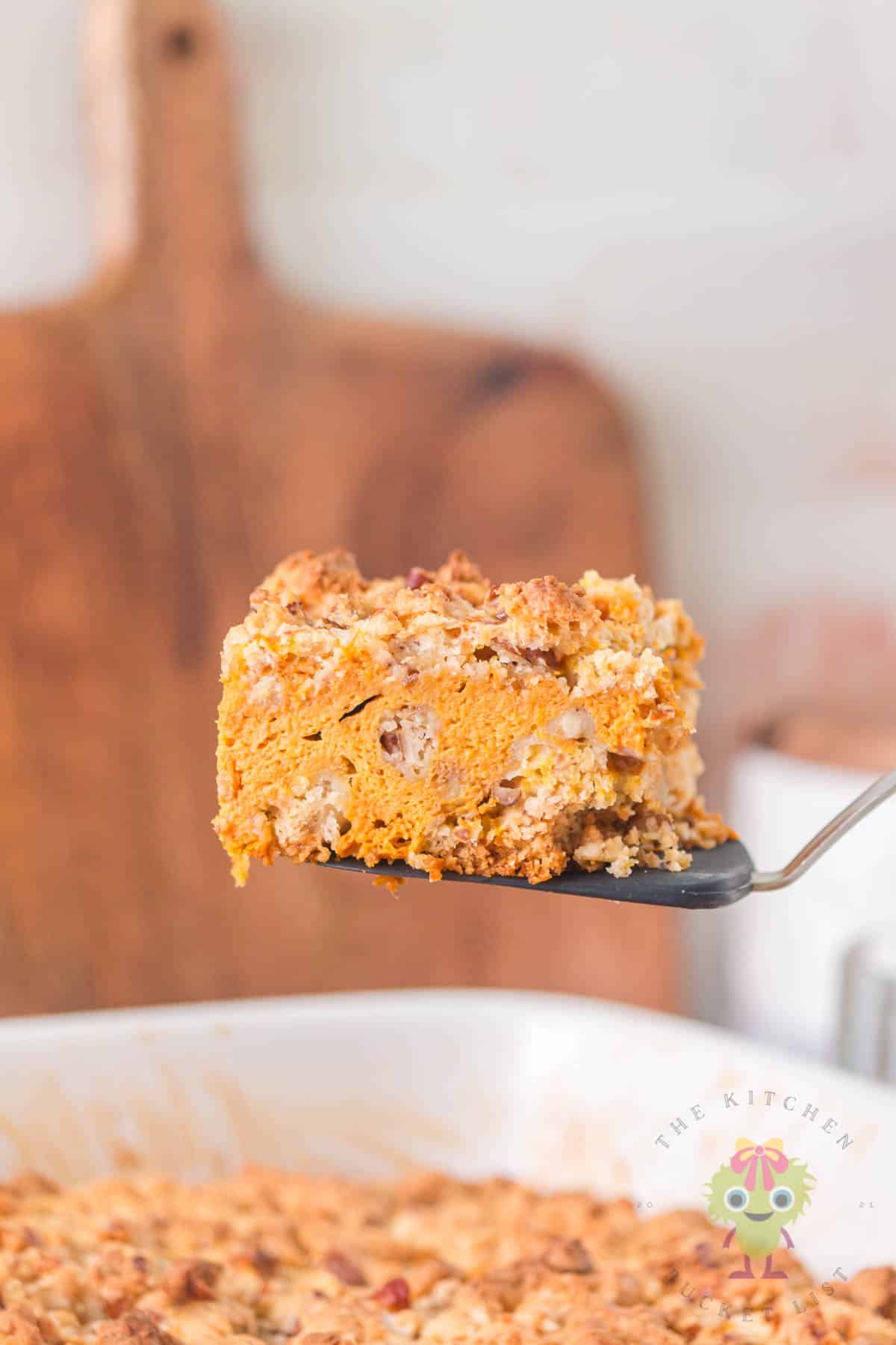 A piece of pumpkin pie cake with its warm pumpkin spice creamy pie-like filling and crunchy golden buttery topping. It is being served from the 9x13 dish, which makes it the perfect make-ahead cozy fall dessert.