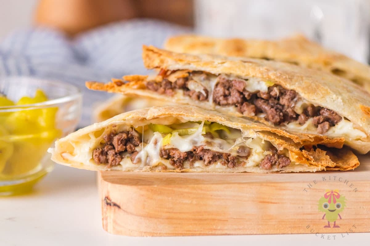Close-up of sliced air fryer Mississippi ground beef quesadillas on a wooden board with gooey melted cheese pull and pepperoncini in the filling. Small bowl of pepperoncini in the background.