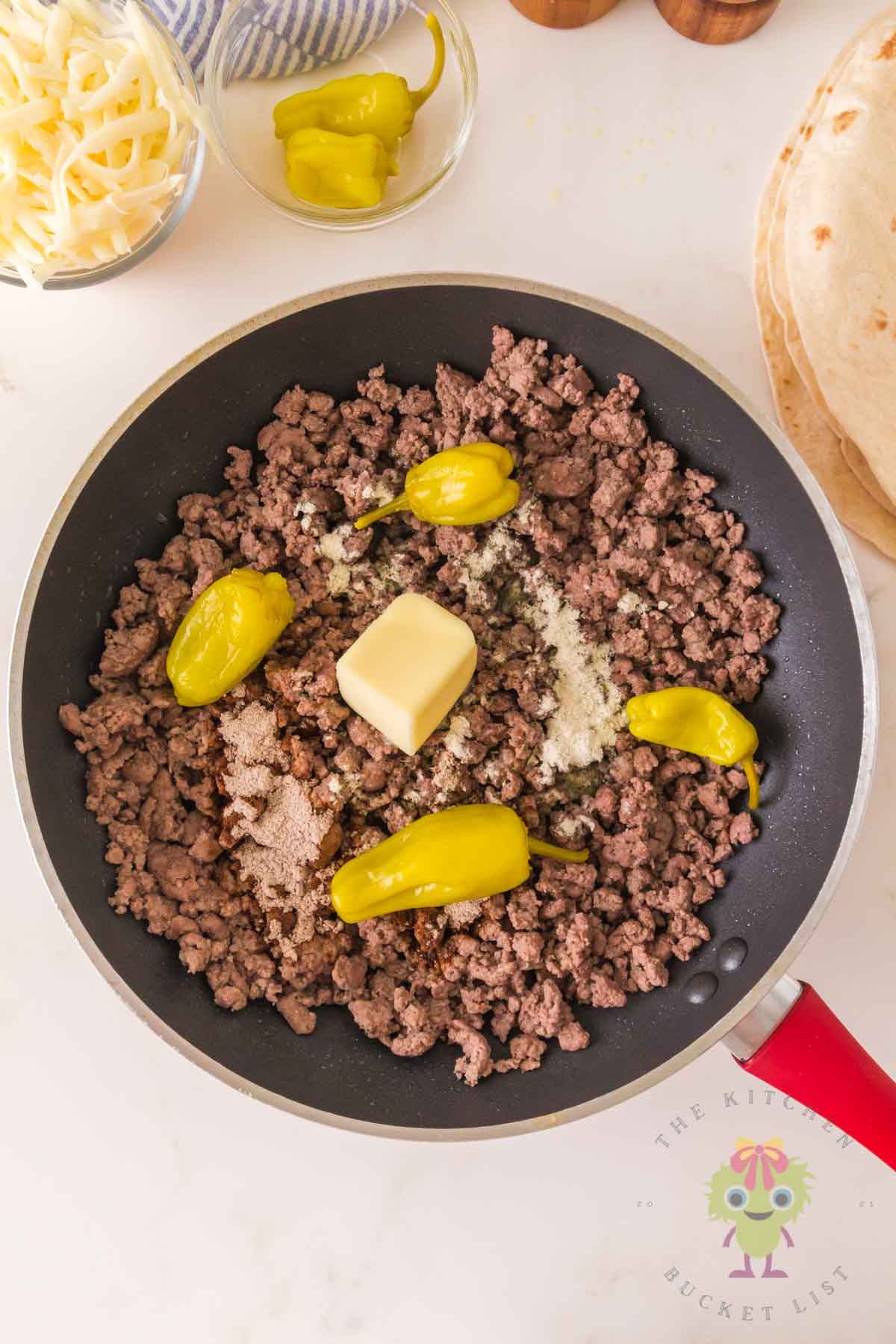 Ground beef cooking in a skillet with a pat of butter, ranch seasoning, au jus mix, and whole pepperoncini peppers on top.